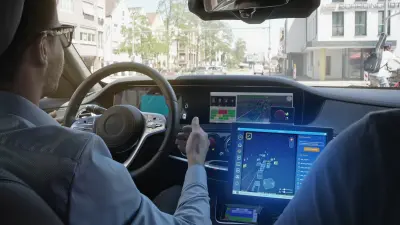 A man sits in the driver’s seat of a test vehicle for autonomous driving. He keeps his hands close to the steering wheel without touching it. The cockpit contains several screens that display the environment as perceived by the various sensors.
