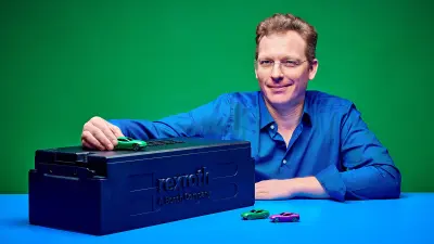 Ein Mann in blauem Hemd, der neben einem schwarzen Batteriemodul mit Bosch-Rexroth-Aufdruck und drei Spielzeug-E-Autos sitzt.
