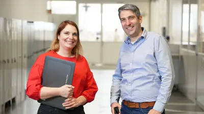 Julia ist links und trägt eine rote Bluse, sie hat ihren Laptop im rechten Arm vor sich und lächelt in die Kamera. Christoph steht rechts daneben, er trägt ein blaues Hemd, seine Hände ruhen auf seiner Hüfte, in der linken trägt er ein Handy und er lächelt in die Kamera.