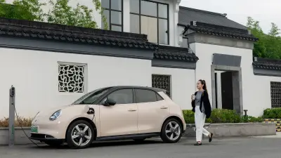A woman leans on a car with a smartphone in her hand. She is watching a man plug a charging cable into the car’s charging port.