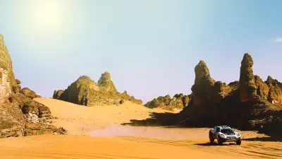 One of the racing SUVs drives through a desert and churns up sand. The sun shines in the sky.