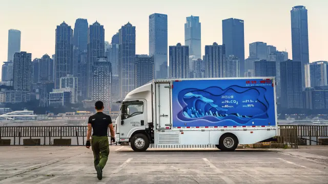 Ein junger Mann im schwarzen T-Shirt geht, von hinten gesehen, auf einen quer stehenden blau-weißen Lastwagen zu. Am Horizont die Skyline einer Millionenstadt.
