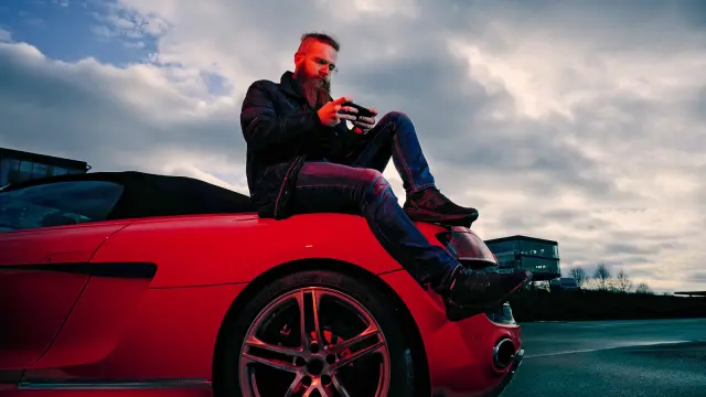 Florian Roschu, world champion in the smartphone racing game "Asphalt 9", sits on the road and leans against a red sports car while playing on his smartphone.
