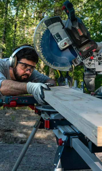 Der Holzkünstler Adam Detre steht umgeben von Bäumen hinter einer kabellosen Kapp- und Gehrungssäge Bosch GCM 18V-305 GDC Professional und richtet ein langes Holzstück sorgfältig aus.