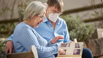 Clemens Weller and his mother Ute Weller sit on a park bench and look at a photo album. He wears a mask.