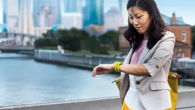 A person stands near a waterfront, the background shows an urban cityscape with tall buildings and a bridge.