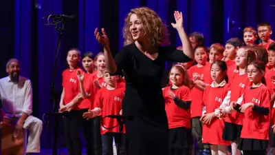 Person conducting a children’s choir on stage, microphone in front, blue backdrop, red shirts.